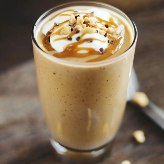 A creamy keto peanut butter smoothie in a glass topped with crushed peanuts and a straw, placed on a light brown table.
